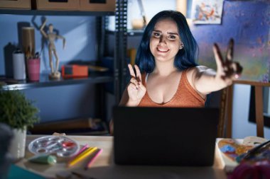Young modern girl with blue hair sitting at art studio with laptop at night smiling looking to the camera showing fingers doing victory sign. number two. 