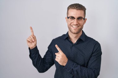 Young caucasian man standing over isolated background smiling and looking at the camera pointing with two hands and fingers to the side. 