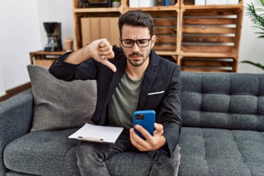 Young hispanic psychologist man doing therapy on video call with smartphone with angry face, negative sign showing dislike with thumbs down, rejection concept 