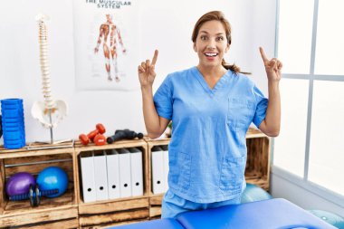 Middle age hispanic physiotherapist woman working at pain recovery clinic smiling amazed and surprised and pointing up with fingers and raised arms. 