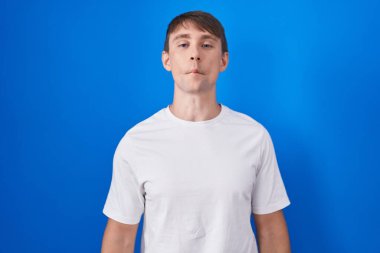 Caucasian blond man standing over blue background making fish face with lips, crazy and comical gesture. funny expression. 