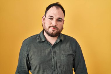 Plus size hispanic man with beard standing over yellow background relaxed with serious expression on face. simple and natural looking at the camera. 