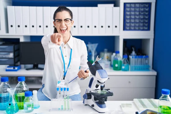 Young brunette woman working at scientist laboratory pointing displeased and frustrated to the camera, angry and furious with you