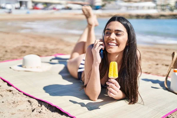 若いですヒスパニック女性話す上のスマートフォン食べるアイスクリーム嘘砂の上に海辺 — ストック写真