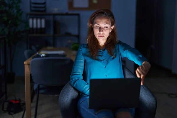 Brunette woman working at the office at night looking unhappy and angry showing rejection and negative with thumbs down gesture. bad expression.
