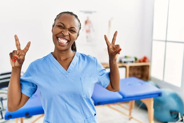 Black woman with braids working at pain recovery clinic smiling with tongue out showing fingers of both hands doing victory sign. number two.