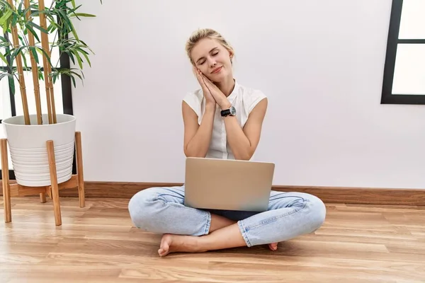 Young blonde woman using computer laptop sitting on the floor at the living room sleeping tired dreaming and posing with hands together while smiling with closed eyes.