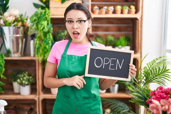 Hispanic young woman working at florist holding open sign scared and amazed with open mouth for surprise, disbelief face