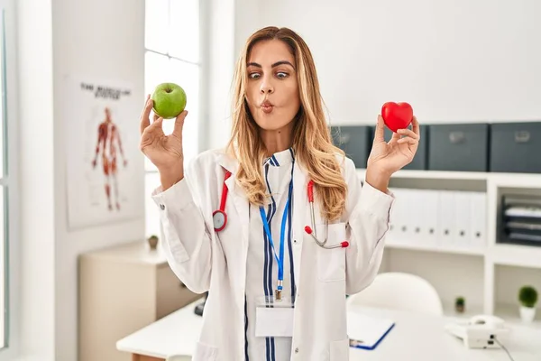 Young blonde doctor woman holding heart and green apple making fish face with mouth and squinting eyes, crazy and comical.