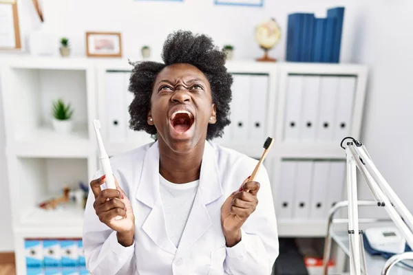 African dentist woman holding electric toothbrush and normal toothbrush angry and mad screaming frustrated and furious, shouting with anger looking up.