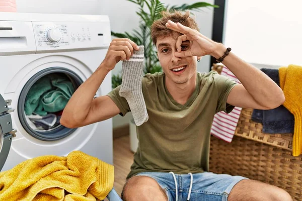 Young caucasian man doing laundry holding sock smiling happy doing ok sign with hand on eye looking through fingers