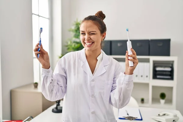 Young Hispanic Dentist Woman Holding Ordinary Toothbrush Electric Toothbrush Winking — Stok Foto