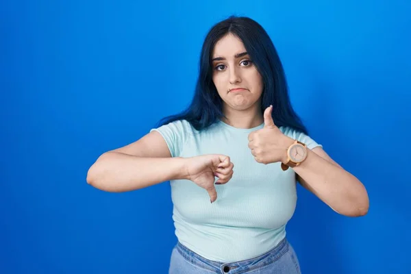 Young modern girl with blue hair standing over blue background doing thumbs up and down, disagreement and agreement expression. crazy conflict