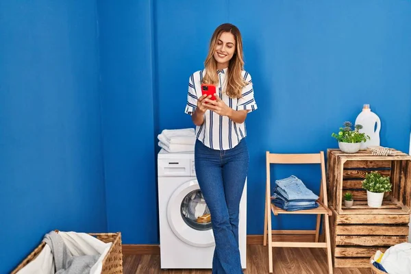 洗濯室で洗濯機を待っているスマートフォンを使用して若いブロンドの女性 — ストック写真