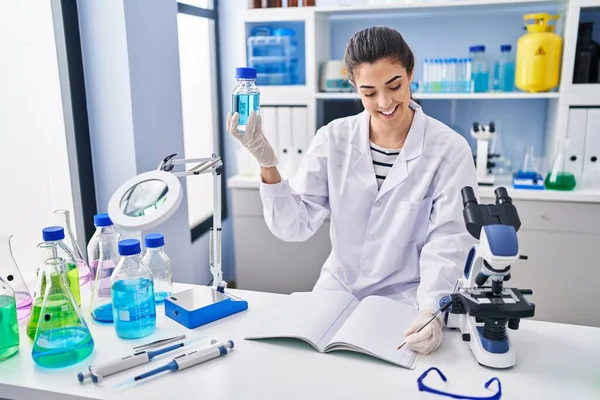 Young beautiful hispanic woman scientist holding bottle writing on notebook at laboratory