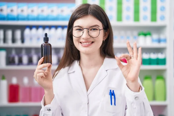 若い白人女性は薬局で働いています美容液を保持します指でOkサインを行います 笑顔優しいジェスチャー優れたシンボル — ストック写真