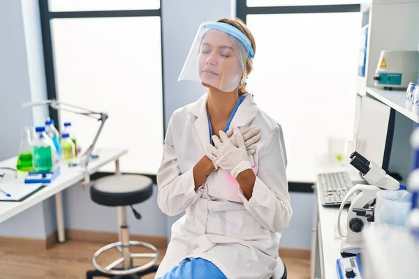 在科学家实验室工作的金发年轻女人戴着面具 双手放在胸前笑着 满脸感激地闭上眼睛 健康概念 — 图库照片