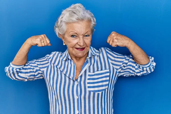 头发灰白的老妇人站在蓝色的背景上 露出胳膊肌肉 露出自豪的笑容 健身概念 — 图库照片
