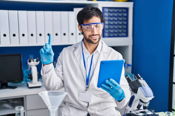 Handsome latin man working at scientist laboratory using tablet smiling with an idea or question pointing finger with happy face, number one 