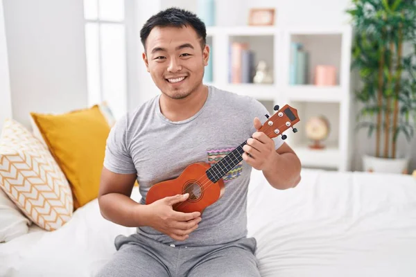 Young chinese man playing ukulele sitting on bed at bedroom