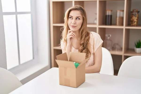 Beautiful blonde woman with cardboard box thinking concentrated about doubt with finger on chin and looking up wondering