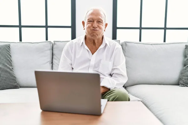 老人在家里用笔记本电脑 坐在沙发上 两颊张着滑稽的脸 气喘吁吁的嘴 疯狂的表情 — 图库照片