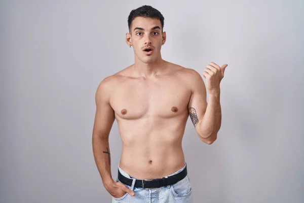 Handsome hispanic man standing shirtless surprised pointing with hand finger to the side, open mouth amazed expression.