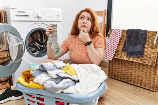 若い赤毛の女性が洗濯機に汚れた洗濯機を入れて質問を心配し 心配し 顎に手で緊張 — ストック写真