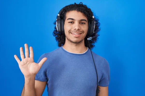 头发卷曲的西班牙裔男人一边微笑着自信而快乐地听着音乐 一边用耳机显示和指指5号 — 图库照片
