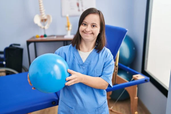 Down syndrome woman wearing physiotherapy uniform holding ball at physiotherapist clinic