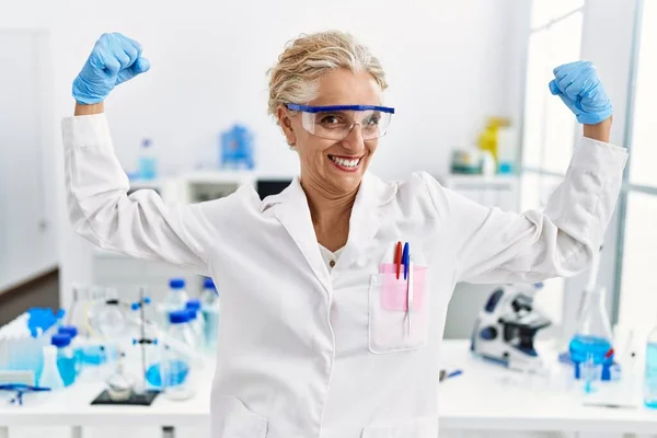 在科学家实验室工作的中年金发女人露出臂膀肌肉 露出自豪的笑容 健身概念 — 图库照片