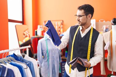 Young hispanic man tailor holding cloths at atelier