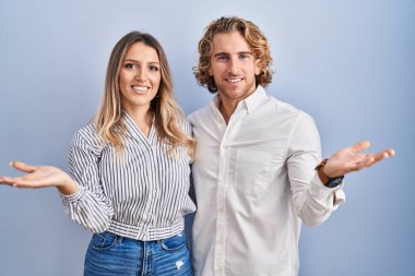 Young couple standing over blue background smiling cheerful presenting and pointing with palm of hand looking at the camera. 