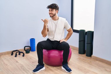 Sakallı İspanyol bir adam yoga odasında pilatese oturmuş gülümsüyor, gülümsüyor ve başparmağını kaldırarak yüzünü işaret ediyor.. 