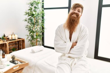 Young redhead man wearing bathrobe sitting on massage table at beauty center