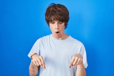 Mavi arka planda dikilen Latin asıllı genç adam parmaklarını göstererek reklamını yapıyor, şaşırmış yüz ve ağzını açıyor. 