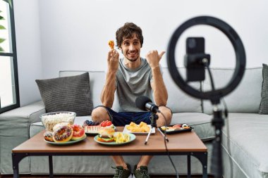 Genç, İspanyol asıllı bir adam akıllı telefonuyla yemek videosu kaydediyor. Başparmağıyla yan tarafa bakıyor ve ağzı açık gülümsüyor. 