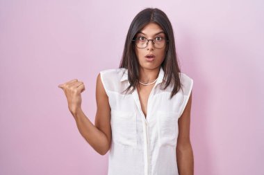 Brunette young woman standing over pink background wearing glasses surprised pointing with hand finger to the side, open mouth amazed expression. 