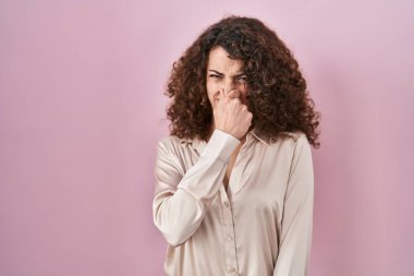 Hispanic woman with curly hair standing over pink background smelling something stinky and disgusting, intolerable smell, holding breath with fingers on nose. bad smell 