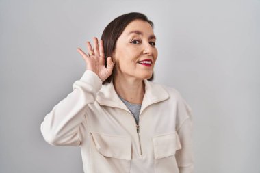 Middle age hispanic woman standing over isolated background smiling with hand over ear listening an hearing to rumor or gossip. deafness concept. 