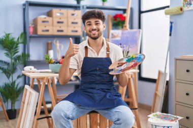 Arab man with beard painter sitting at art studio holding palette smiling happy and positive, thumb up doing excellent and approval sign 