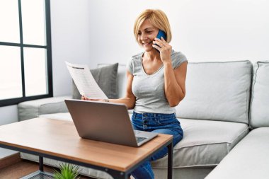 Orta yaşlı sarışın kadın evde dizüstü bilgisayar kullanarak akıllı telefondan konuşuyor.