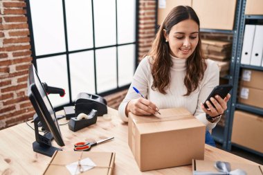Genç ve güzel İspanyol kadın ecommerce iş işçisi. Ofisteki pakette akıllı telefon yazısını kullanıyor.