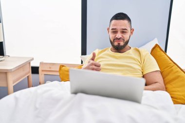 Hispanic man using laptop on the bed smiling happy and positive, thumb up doing excellent and approval sign 