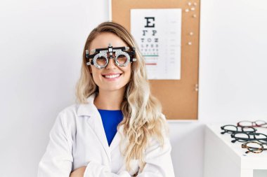 Klinikte optometri gözlüğü takan, kollarını kavuşturup kameraya gülümseyen genç ve güzel bir gözlükçü kadın. pozitif kişi. 