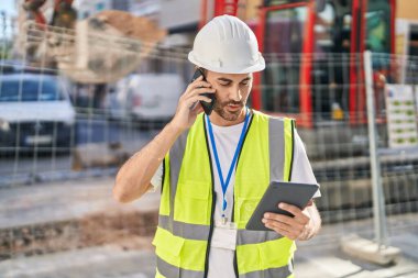 Young hispanic man architect talking on smartphone using touchpad at street