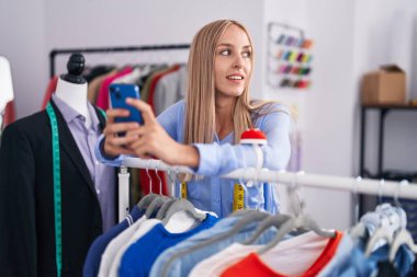 Young blonde woman tailor using smartphone leaning on rack at tailor shop