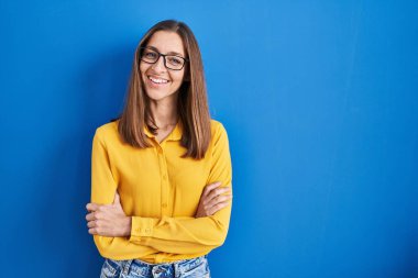 Young woman wearing glasses standing over blue background happy face smiling with crossed arms looking at the camera. positive person. 