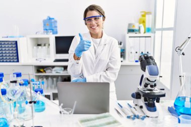 Young hispanic woman wearing scientist uniform working at laboratory smiling happy and positive, thumb up doing excellent and approval sign 
