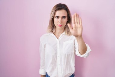Young beautiful woman standing over pink background doing stop sing with palm of the hand. warning expression with negative and serious gesture on the face. 